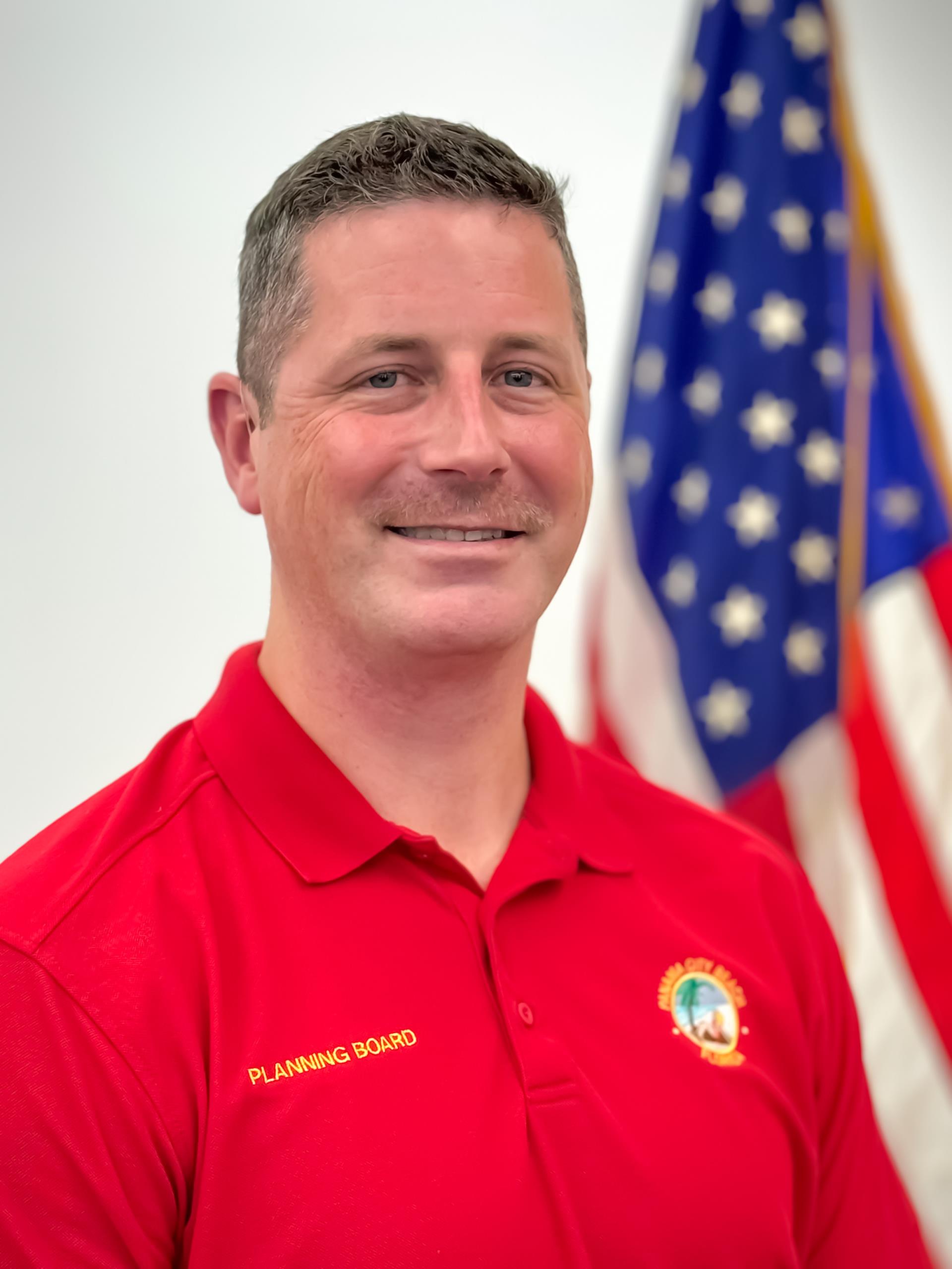 Planning Board Member, Zachary Coleman wearing a red shirt standing in front of American flag