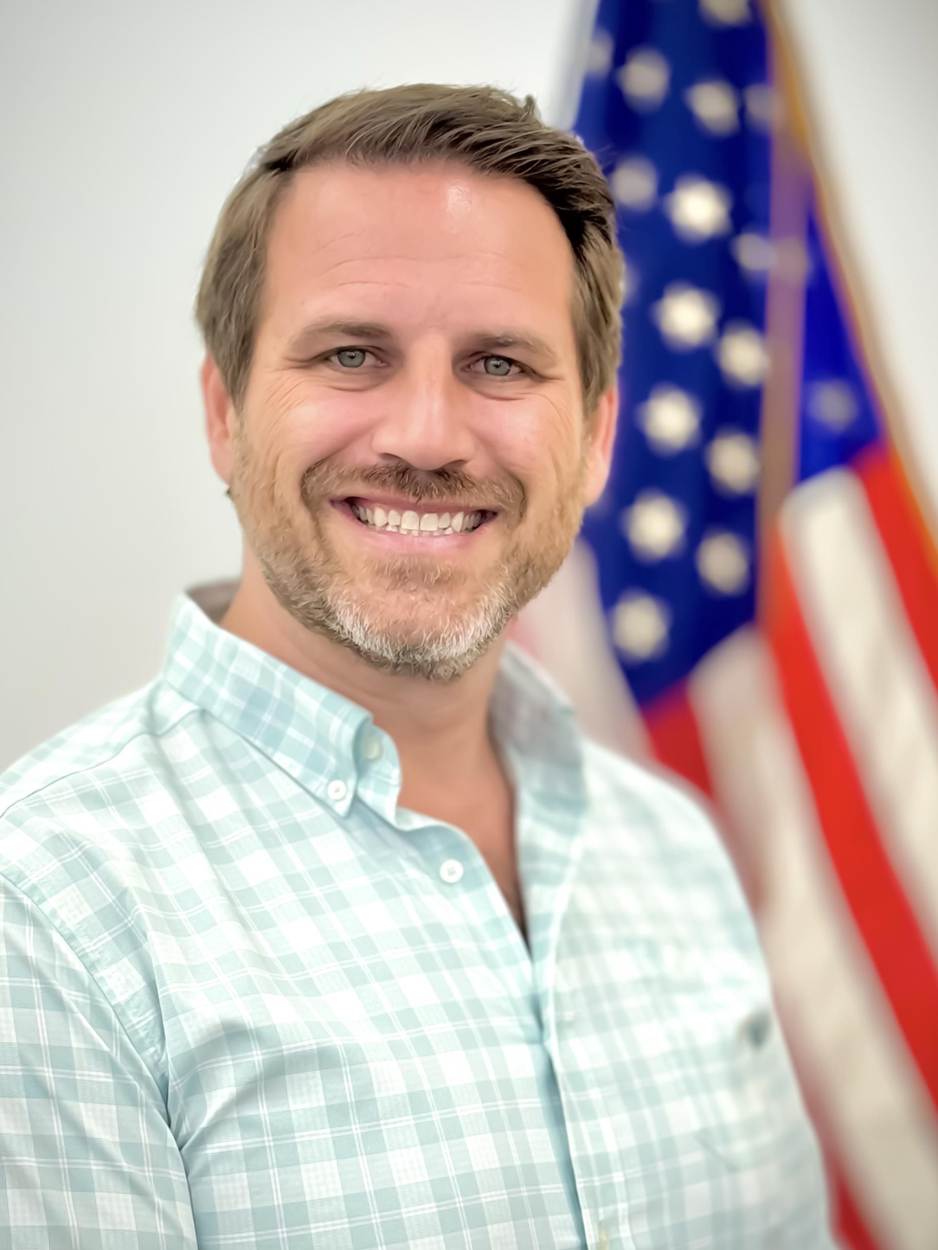 Planning Board Chairman, Josh Wakstein wearing a white and aqua shirt standing in front of American flag