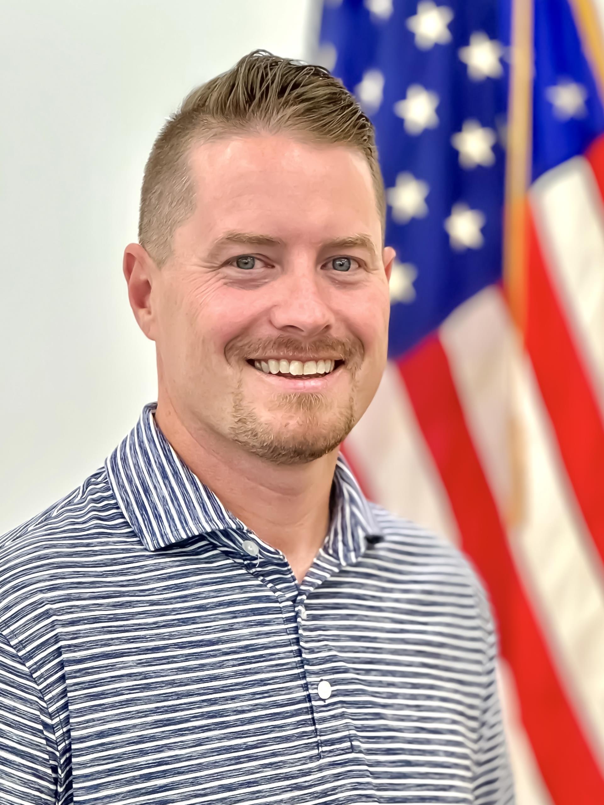 Planning Board Member, Grant Wittstruck wearing a navy blue and white shirt standing in front of American flag